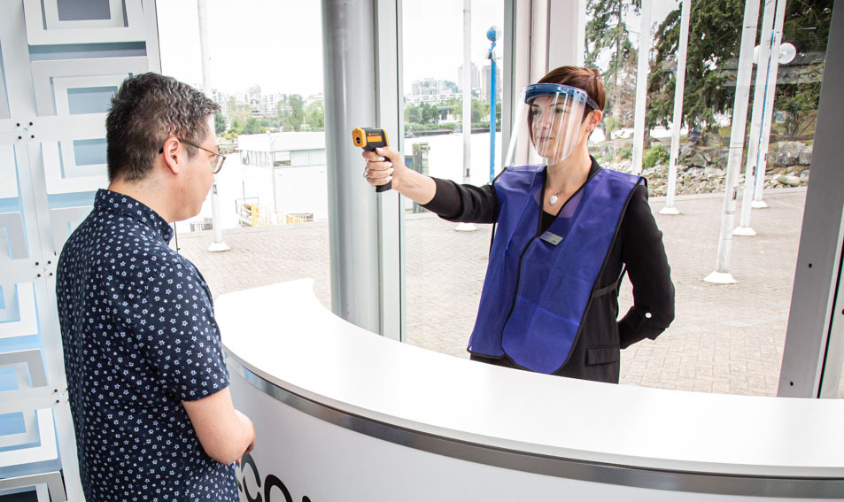 A COVID Safety Ambassadorâ„¢ wearing an Axiom Face Shield checks the temperature of a guest using a contactless thermometer.